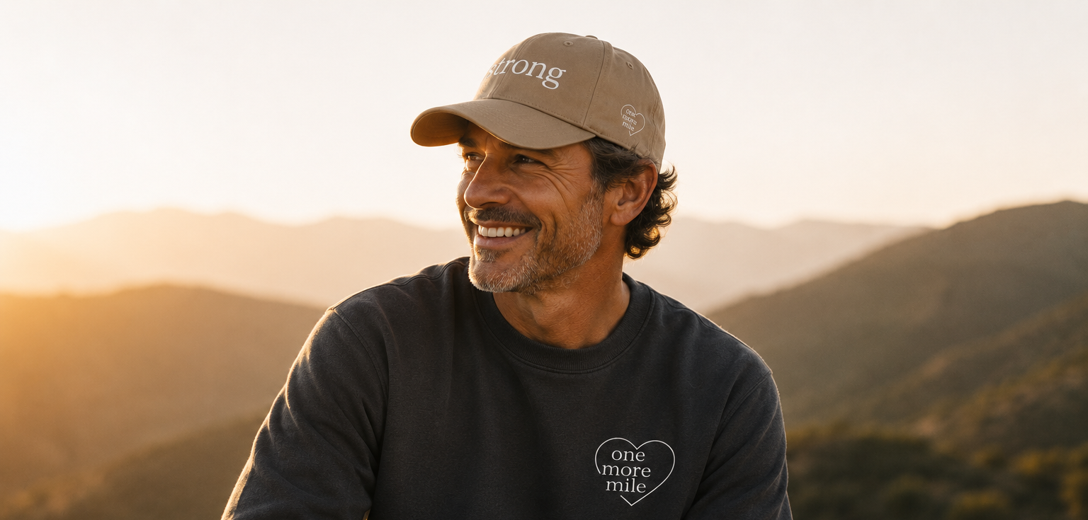 Man wearing a cap and sweatshirt with text, sitting in a mountainous landscape at sunset.