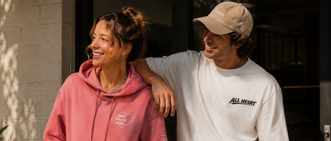 Two people standing together, one wearing a pink hoodie and the other in a white t-shirt with 'All Heart' branding.