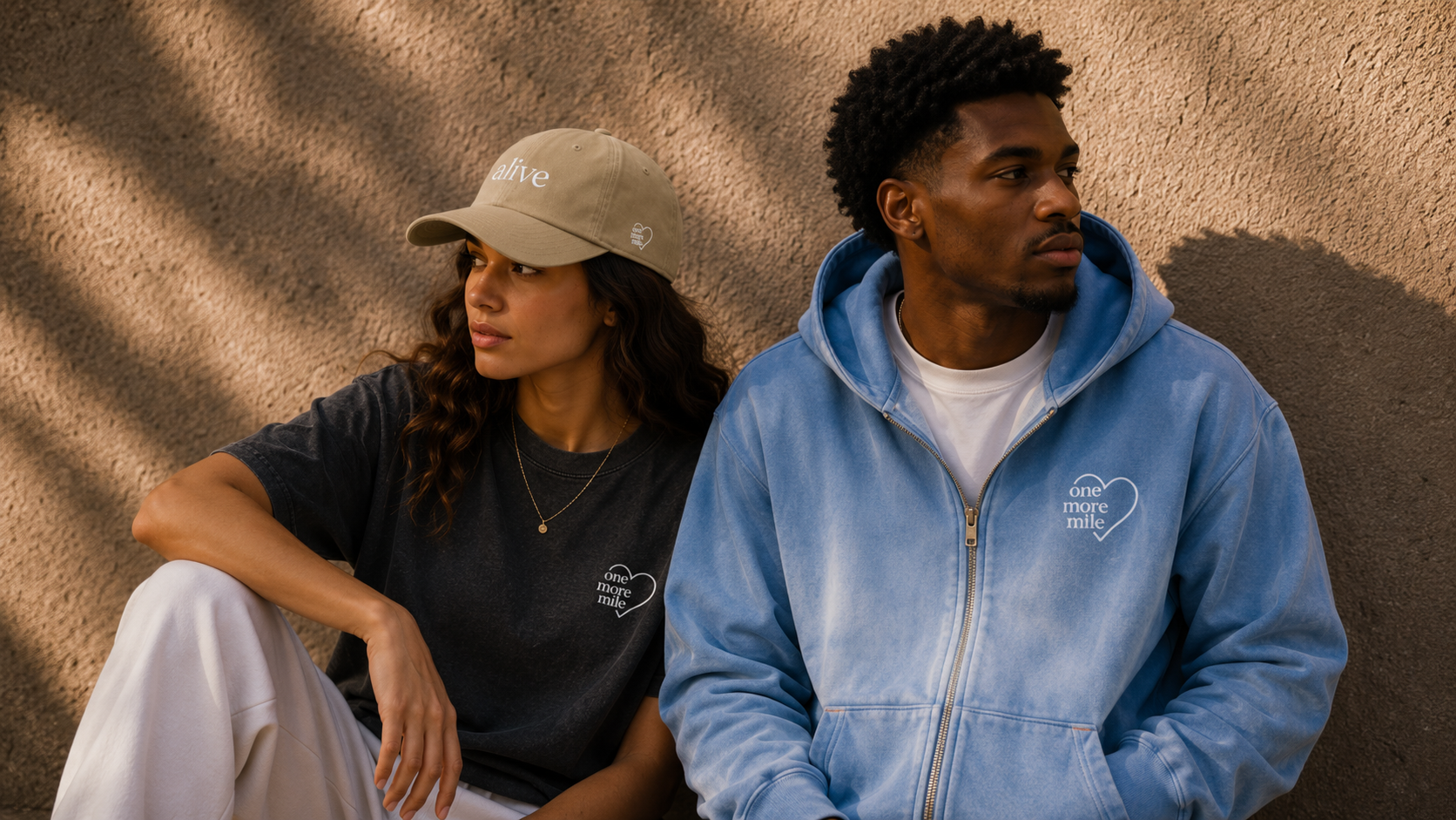 Two people sitting against a beige wall, wearing branded clothing.
